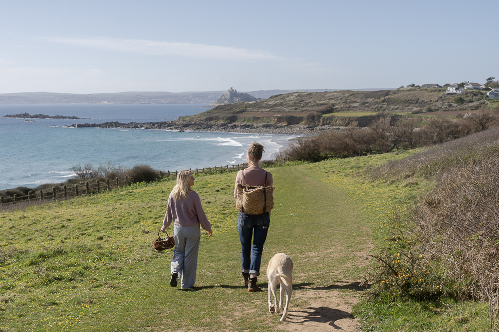 Two women and a dog walking along the coast in Cornwall with foraging baskets