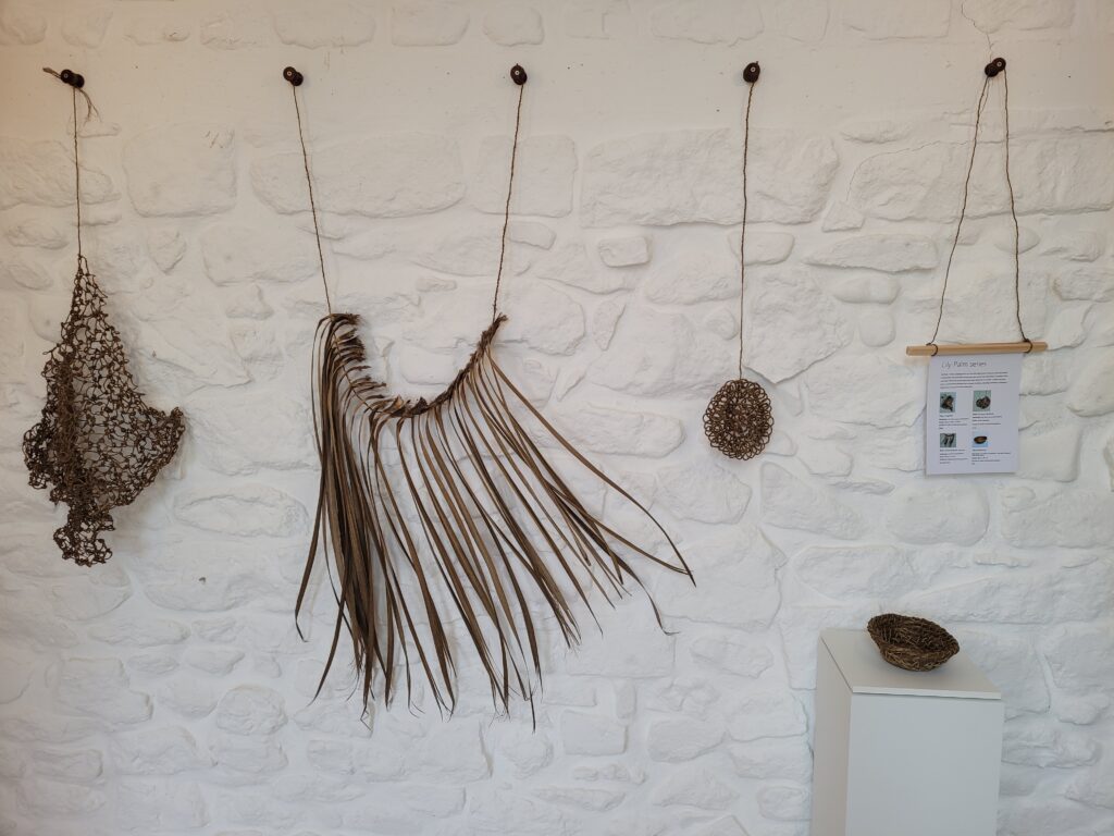 White washed wall with 3 hanging artworks made from cordyline and one bowl on a white stand