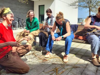 A group of adults sitting outdoors weaving plants with additional plants strewn across the floor