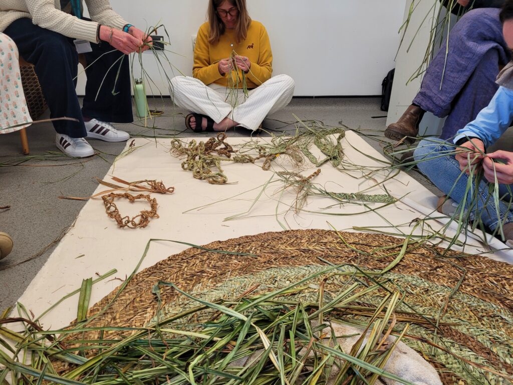People sitting around a canvas sheet with a woven mat, pile of plants and different pieces of plant waving on it