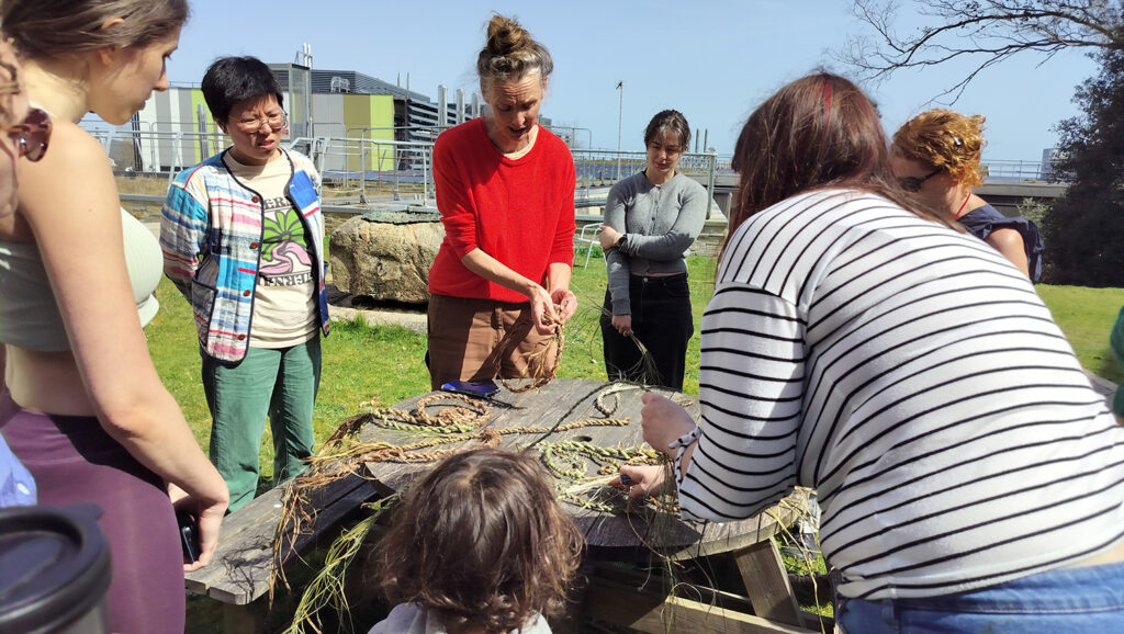 Rachel Lambert introducing a range of natural materials to weave with, standing outdoors with a group of adults