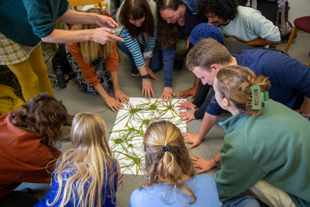 A group sitting around a large white piece of paper with their plant woven creations on it