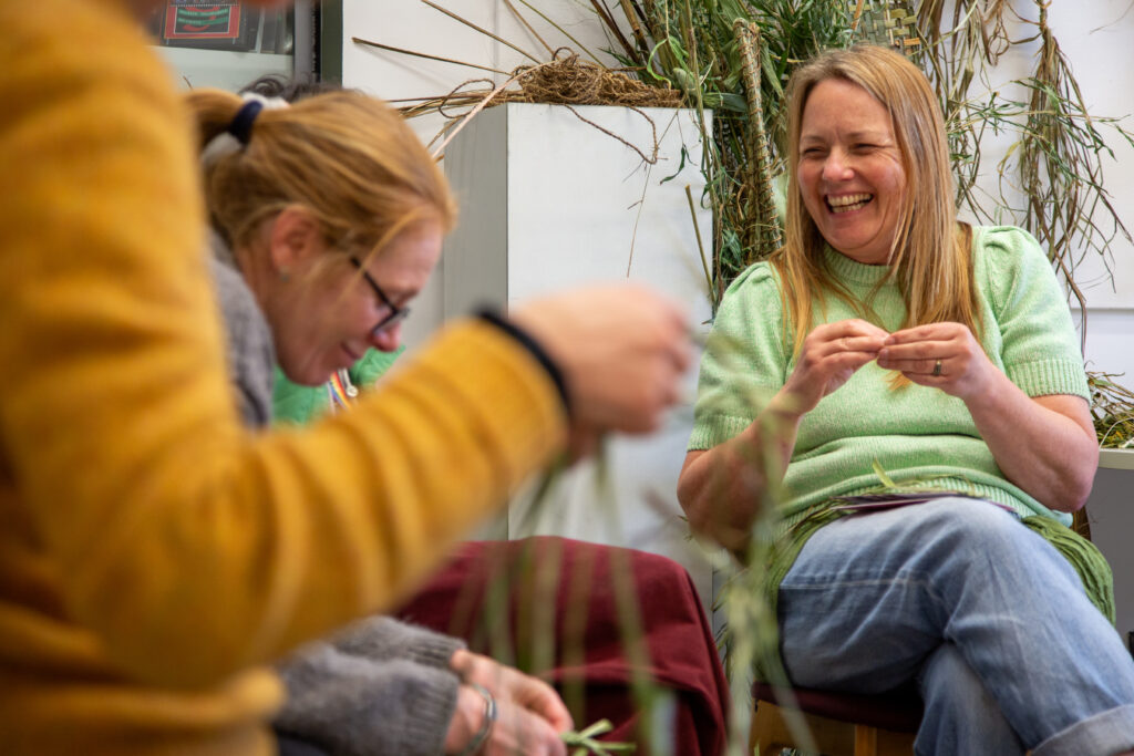 Women laughing and sitting in a room of drying plants creating with the materials around them